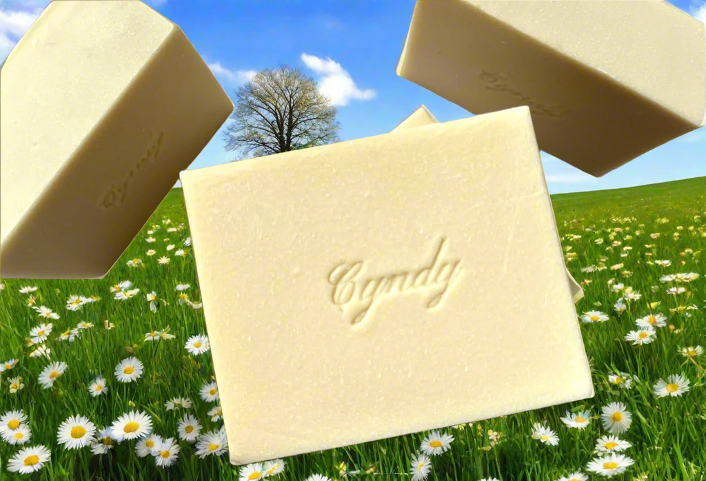 A bar of goat milk soap with the word 'Cyndy' inscribed on it, displayed against a cosmic starry background with a small bird and a Christmas tree in the foreground.