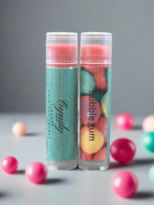 Two lip balm tubes with colorful packaging on a gray surface with scattered bubblegum balls.