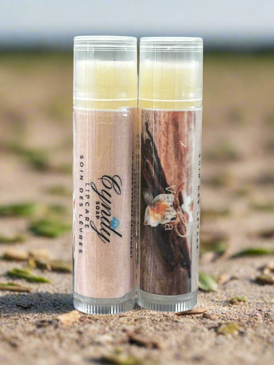 Two lip balm tubes on a sandy surface with a blurred natural background
